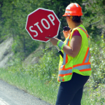 A flagger stands in the road to direct traffic. They're wearing neon and relfective safety gear and speaking into a radio as their left hand holds up a stop sign.