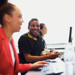 Three students working on computers while talking to their peers sitting at the same desk