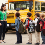 An elementary school teacher smiles at students lining up for the yellow school bus parked in the background