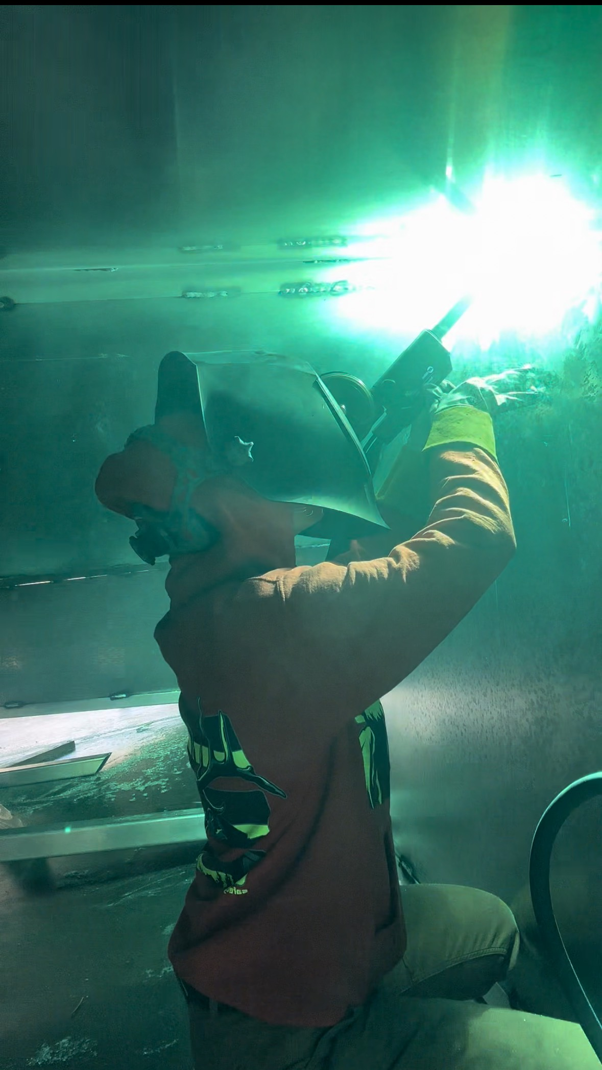 Jessie welding a fixture above their head. The picture is saturated in teal light. 