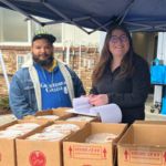 A student and food pantry staff member smile for the camera under the shade of a canopy tent in GHC blue. In the foreground are boxes of food.