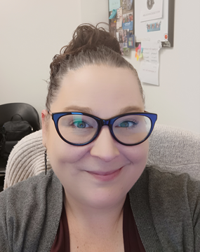 A portrait image of Stephanie Thornton smiling at the camera from her office chair, her curly brown hair is in a bun and she wears dark blue cat eye glasses.