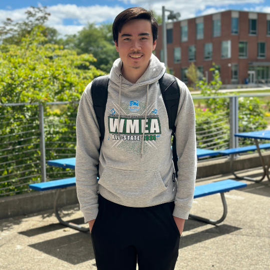 Kevin smiles for the camera, hands in pockets. He's wearing a WMEA hoodie and the GHC campus can be seen in the background