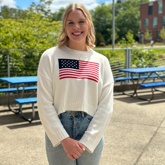 Kristina smiles for the camera, she's wearing an american flag sweater and highwaisted jeans.