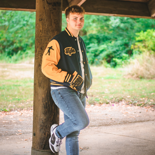 Tyler Poses for the camera against a log beam. He wears a 2024 letterman jacket with his name embroidered on the rightside chest.