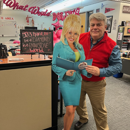 The Aberdeen Campus TRIO office decorated with various pink and Dolly Parton themed balloons. JEB Thorton stands next to a Dolly Parton standie, pretending to hold her book open. On the opposite side of the cut out, is a small chalkboard sign that reads "It's hard to be a Diamond in a Rhinestone World". 