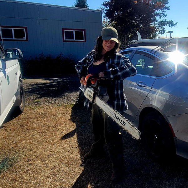 AnnMarie stands between two cars hollding a chainsaw and smiling for the camera.