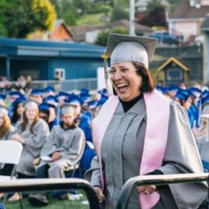 a GED graduate in gray robe and pink sash smiles wide on their way to cross the stage at the commencement ceremony