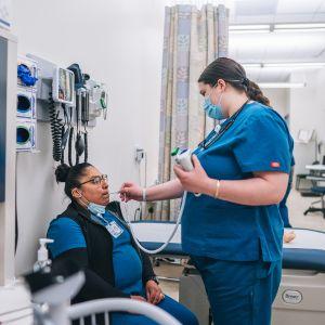 A medical assisting student in blue scrubs, takes the temperature of a peer who sits in a mock up of a doctors office